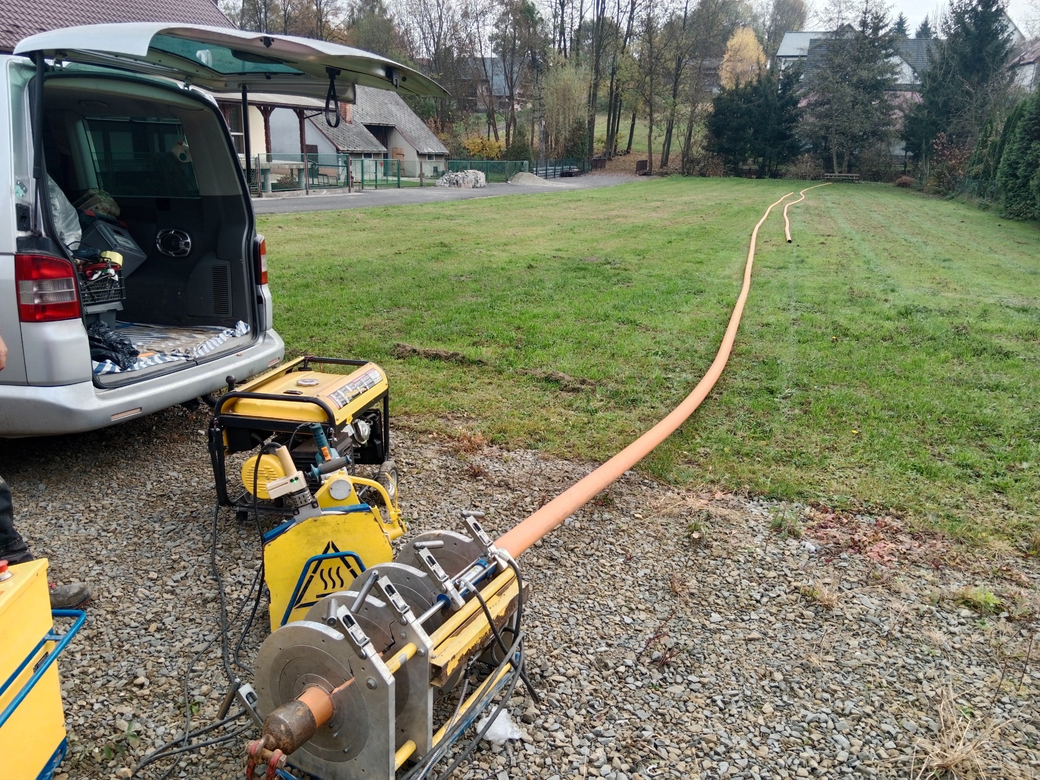 Sprzęt do łączenia rur na żwirowym podłożu. W tle trawnik i zabudowania. Widoczny agregat prądotwórczy i otwarty bagażnik samochodu.