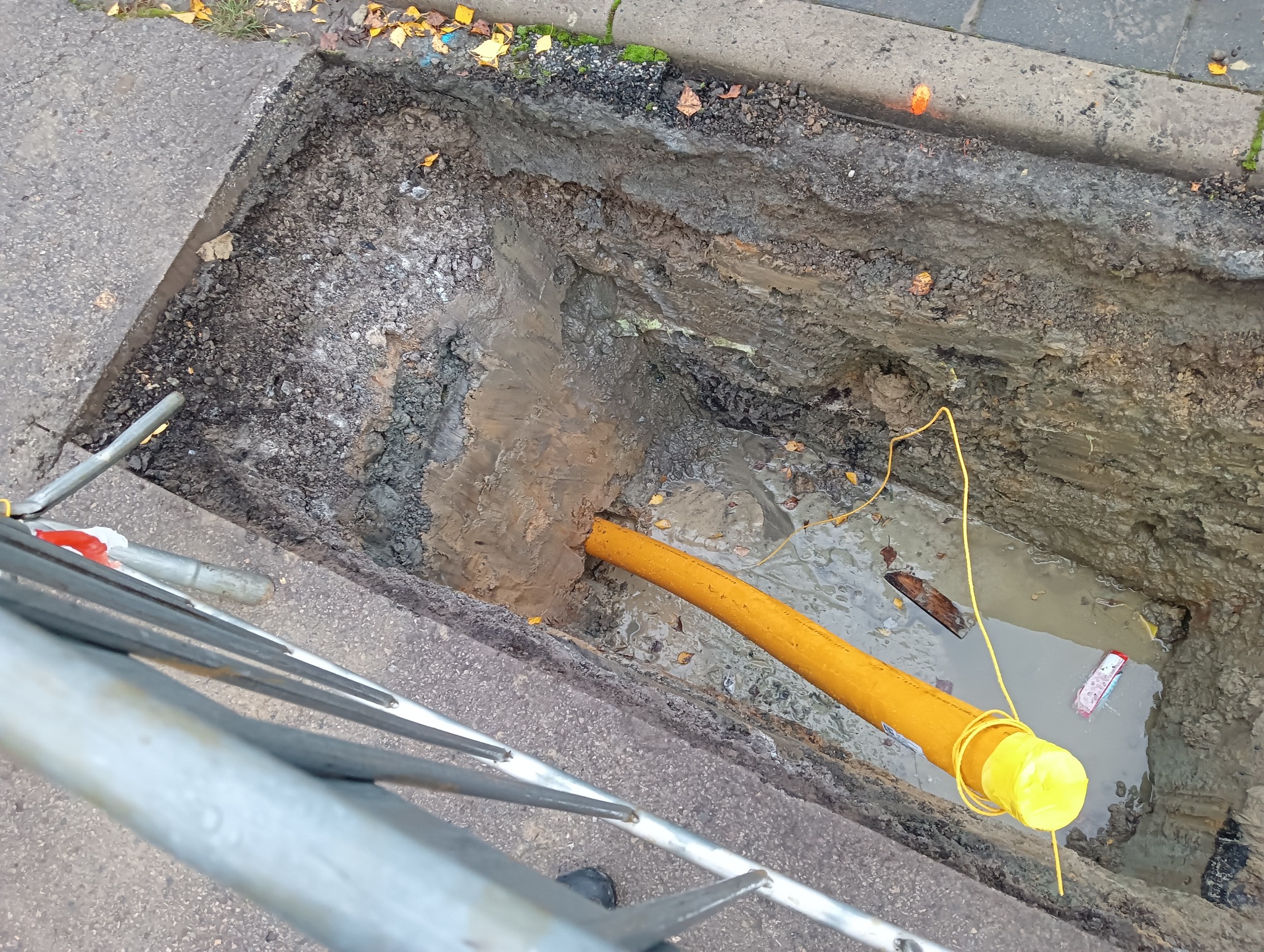 Żółta rura w wykopie ziemnym z wodą, widoczna z góry. Fragment barierek ochronnych w lewym dolnym rogu. Wykop w asfalcie z liśćmi.