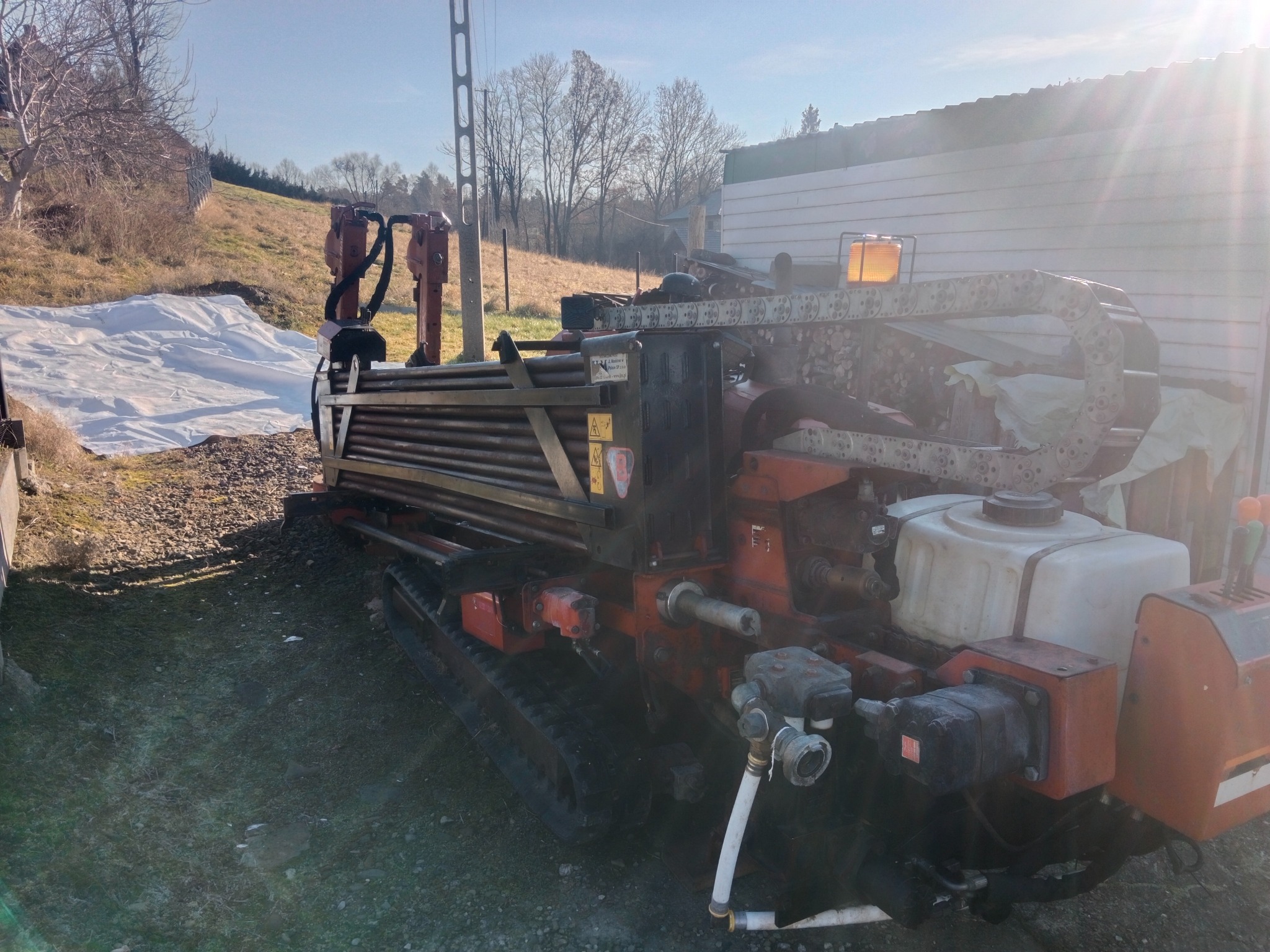 Pomarańczowa maszyna do wiercenia horyzontalnego, z rurami w górnej części, stoi na zewnątrz w słoneczny dzień. Widoczny teren i budynek w tle.