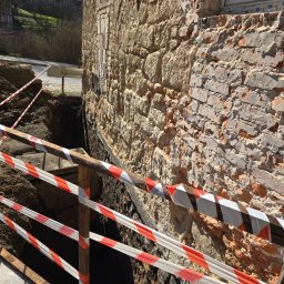 Wykop Roboty Ziemne Krzysztof Jaworski - Wykop zabezpieczony taśmą ostrzegawczą przy fundamencie budynku z kamienia i cegły. Widoczny fragment ulicy i zieleń w tle. Prace ziemne w toku.