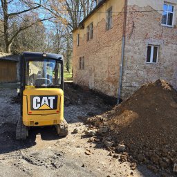 Wykop Roboty Ziemne Krzysztof Jaworski - Żółta mini koparka CAT stoi przed budynkiem z odsłoniętą cegłą i wykopem. Ziemia usypana w pryzmę. Widoczne ślady pracy maszyny.