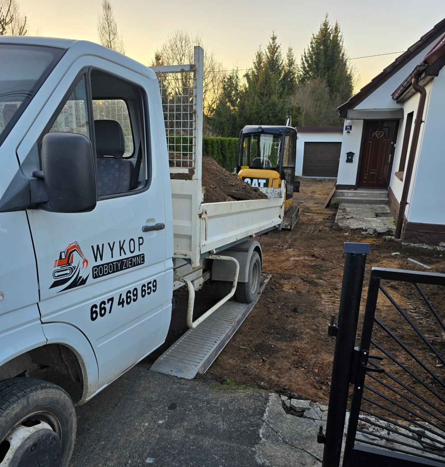 Żółta mini koparka CAT z załadowaną ziemią na przyczepie białego samochodu dostawczego z logo firmy wykonującej roboty ziemne. W tle dom jednorodzinny.