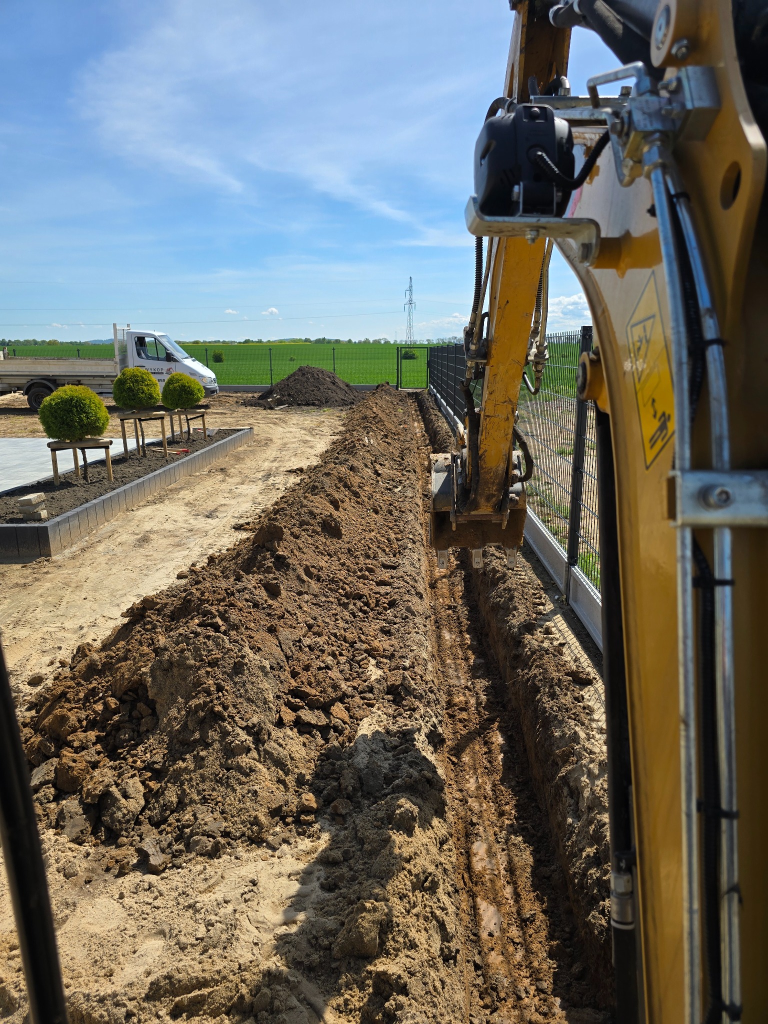 Żółta koparka w trakcie wykopu wąskiego rowu w ziemi, obok ogrodzenie panelowe, w tle trawnik i błękitne niebo. Etap prac ziemnych.
