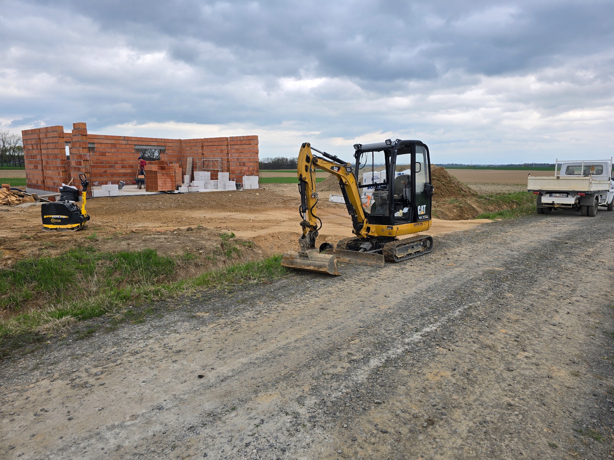 Żółta mini koparka CAT na żwirowej drodze, w tle budowa z czerwonej cegły i biały pick-up. Pochmurne niebo.