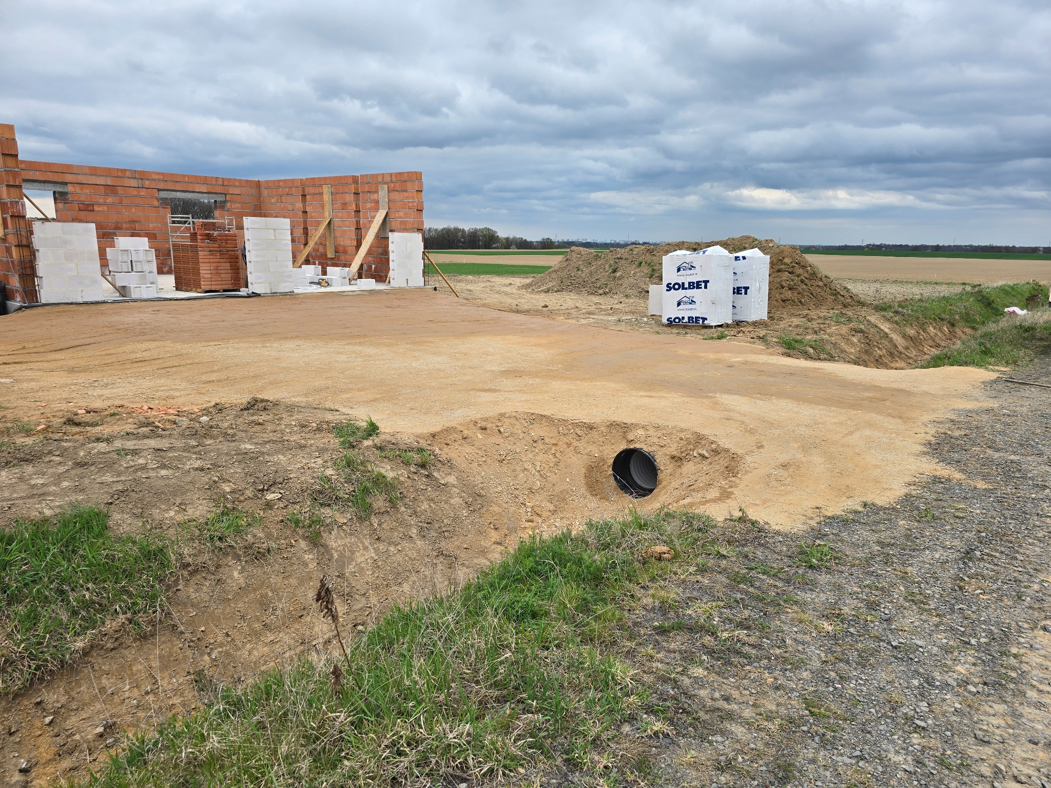 Budowa domu w trakcie: widoczne fundamenty, ściany z cegły, materiały budowlane Solbet i rura drenażowa w wykopie na tle pola i pochmurnego nieba.