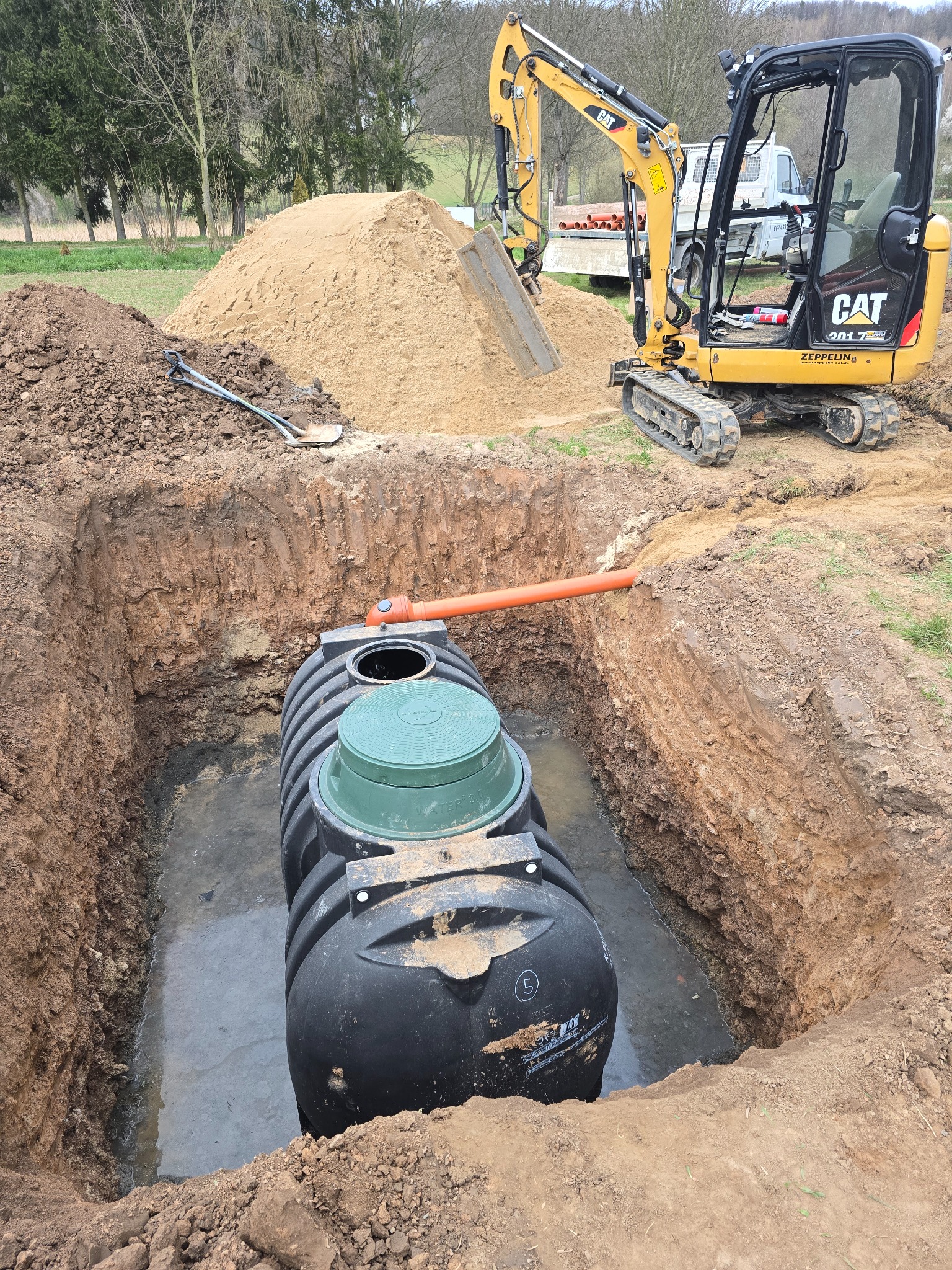 Czarny zbiornik na szambo z zieloną pokrywą, umieszczony w wykopie wypełnionym wodą. W tle koparka CAT i hałda piasku, a obok łopata.