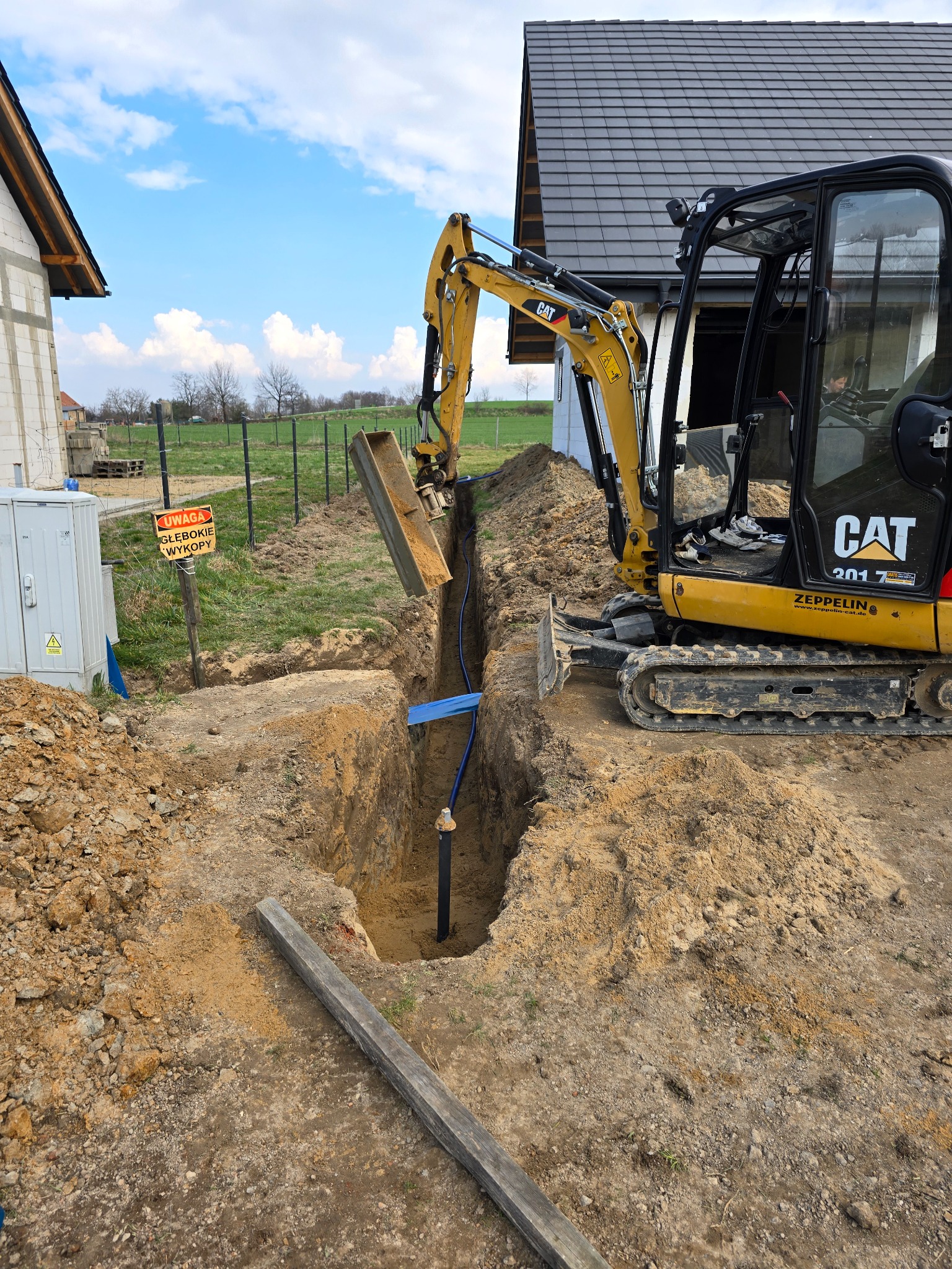 Żółta koparka CAT przy wykopie z rurami na tle budowy domu i zieleni. Tablica ostrzegawcza o głębokich wykopach. Błękitne niebo z chmurami.