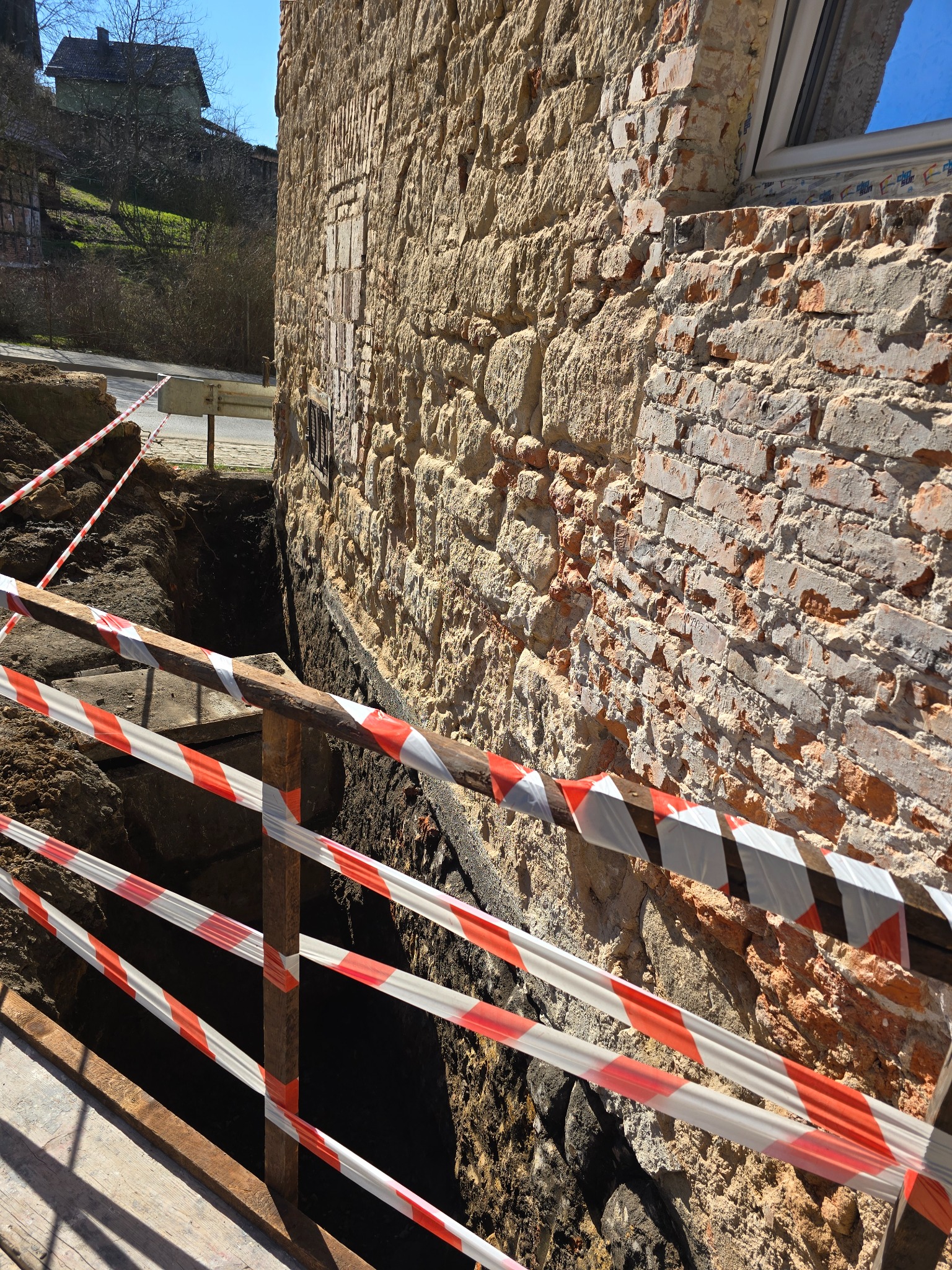 Wykop zabezpieczony taśmą ostrzegawczą przy fundamencie budynku z kamienia i cegły. Widoczny fragment ulicy i zieleń w tle. Prace ziemne w toku.