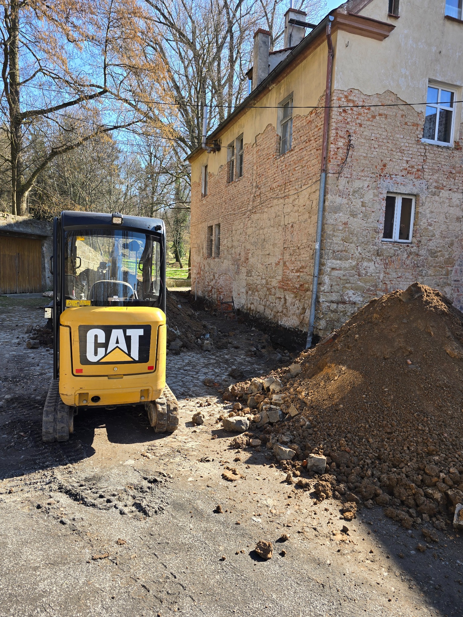 Żółta mini koparka CAT stoi przed budynkiem z odsłoniętą cegłą i wykopem. Ziemia usypana w pryzmę. Widoczne ślady pracy maszyny.
