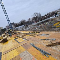 ALSITBUD - Zbrojenie fundamentów budynku na żółtym szalunku, widoczny dźwig i pracownicy w oddali. Praca wre na budowie w pochmurny dzień.