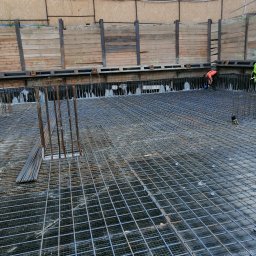 ALSITBUD - Siatka zbrojeniowa na budowie, widoczna z góry. W tle drewniane zabezpieczenia wykopu i pracownik w pomarańczowej koszulce. Widok na zbrojenie fundamentu.