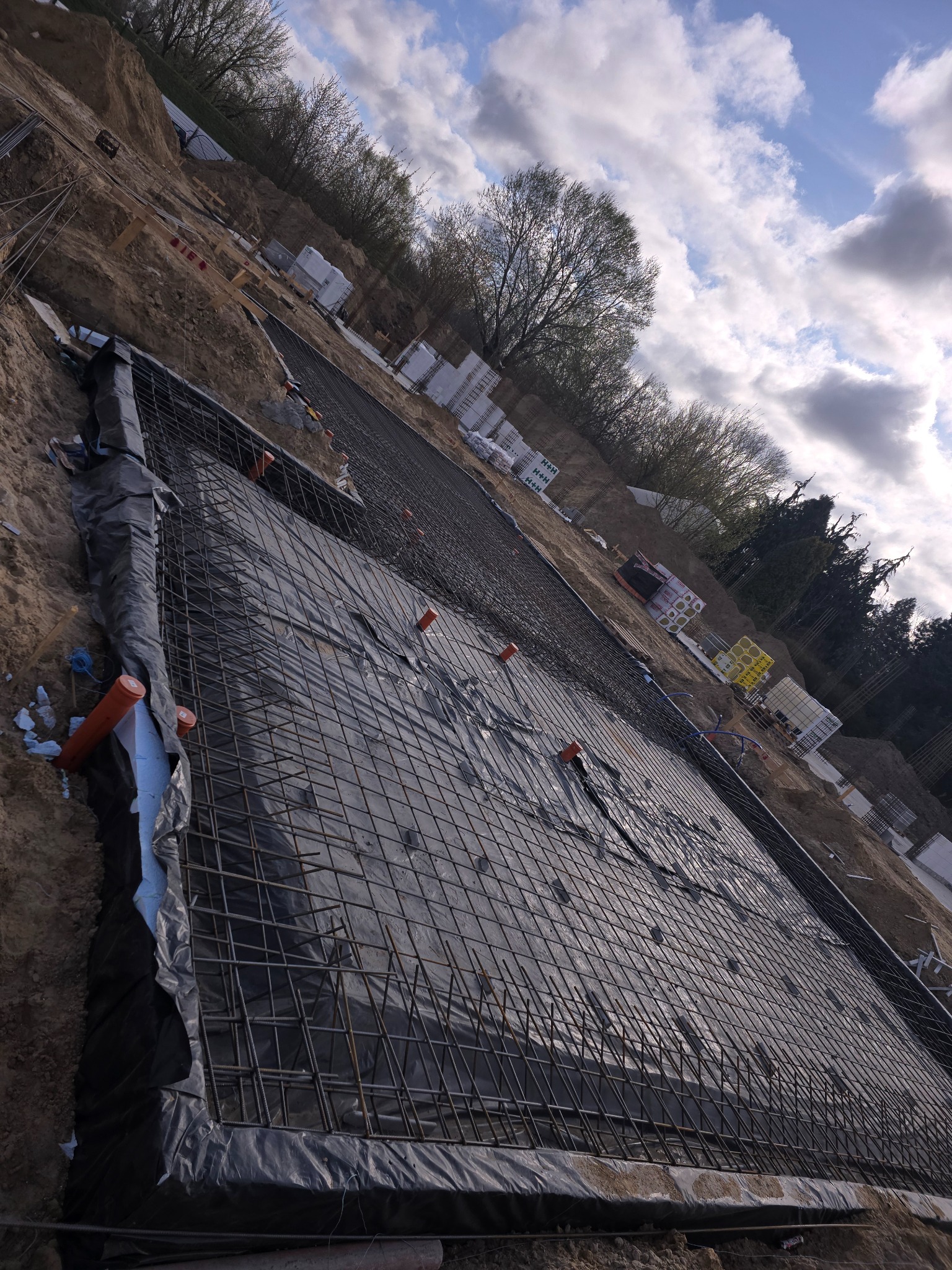 Zbrojenie fundamentów na gruncie z folią i wystającymi rurami. Widok na konstrukcję z prętów stalowych przed zalaniem betonem. W tle materiały budowlane.