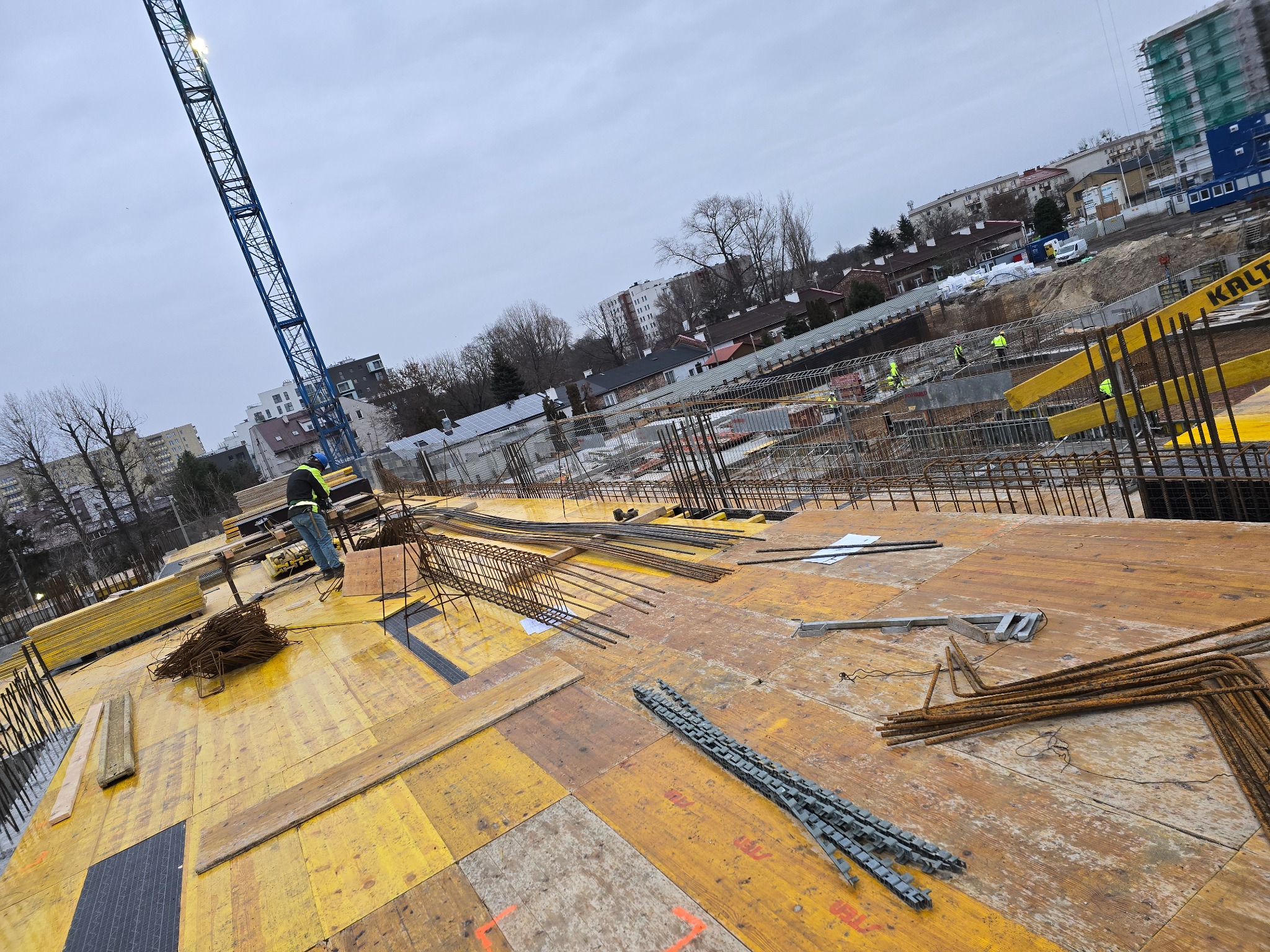 Zbrojenie fundamentów budynku na żółtym szalunku, widoczny dźwig i pracownicy w oddali. Praca wre na budowie w pochmurny dzień.