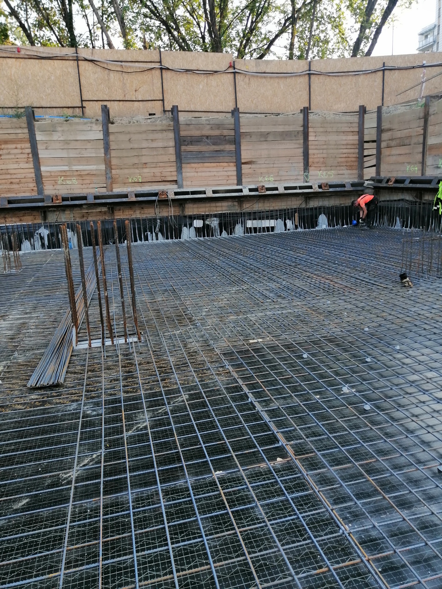 Siatka zbrojeniowa na budowie, widoczna z góry. W tle drewniane zabezpieczenia wykopu i pracownik w pomarańczowej koszulce. Widok na zbrojenie fundamentu.