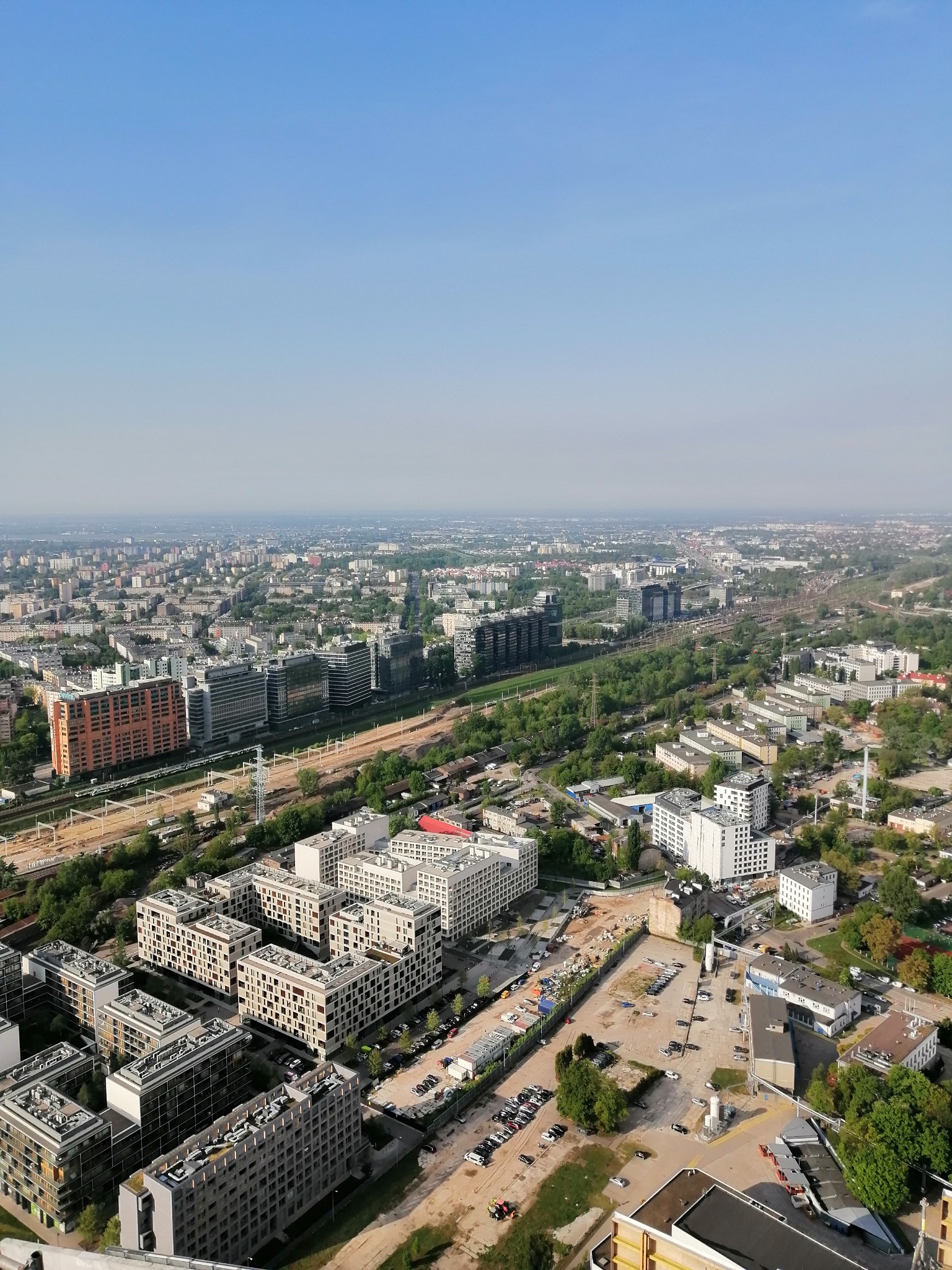Panoramiczny widok z góry na osiedle budynków mieszkalnych w słoneczny dzień, z widoczną infrastrukturą kolejową i rozległą panoramą miasta w tle.