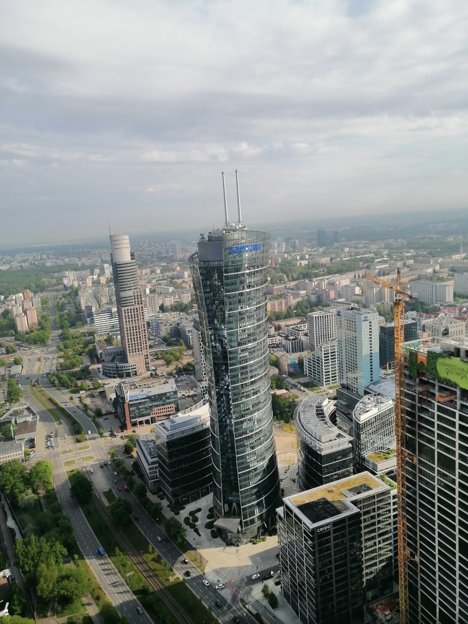 Panorama Warszawy z nowoczesnymi wieżowcami, w tym charakterystyczny szklany budynek z logo Samsung, żuraw budowlany i inne budynki w tle.