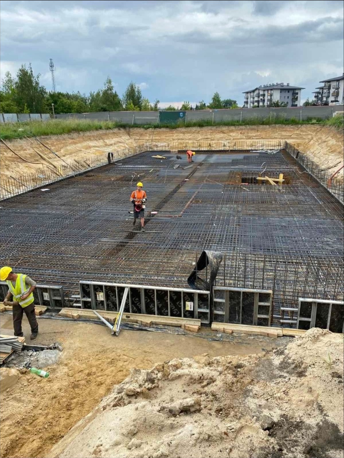 Zbrojenie fundamentów pod budynek wielorodzinny. Dwóch pracowników w kaskach i kamizelkach. Widoczne szalunki i stalowe pręty tworzące siatkę zbrojeniową.