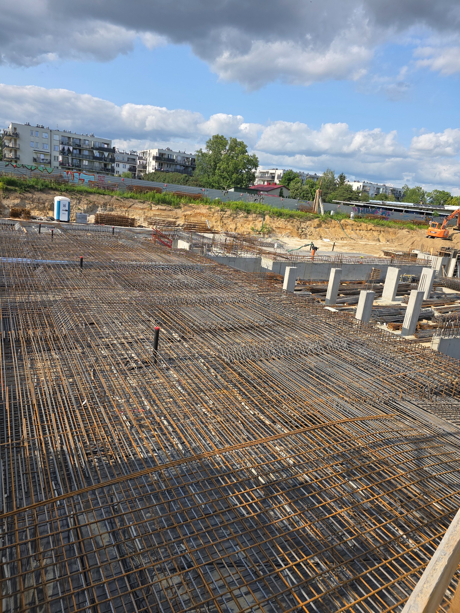Rozległe zbrojenie fundamentów budynku w trakcie budowy, widoczne betonowe podpory, w tle zabudowa miejska i błękitne niebo.