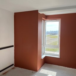 Plytkigladzie - Pokój po malowaniu ścian na kolor terakoty, widoczne białe okno z widokiem na zewnątrz, surowa podłoga i fragment białej ściany z czarnymi listwami.