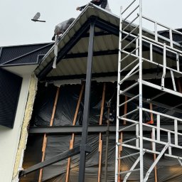 Plytkigladzie - Demontaż fragmentu dachu z rusztowaniem i widoczną konstrukcją ściany z izolacją. Dwóch pracowników na dachu podczas pracy.