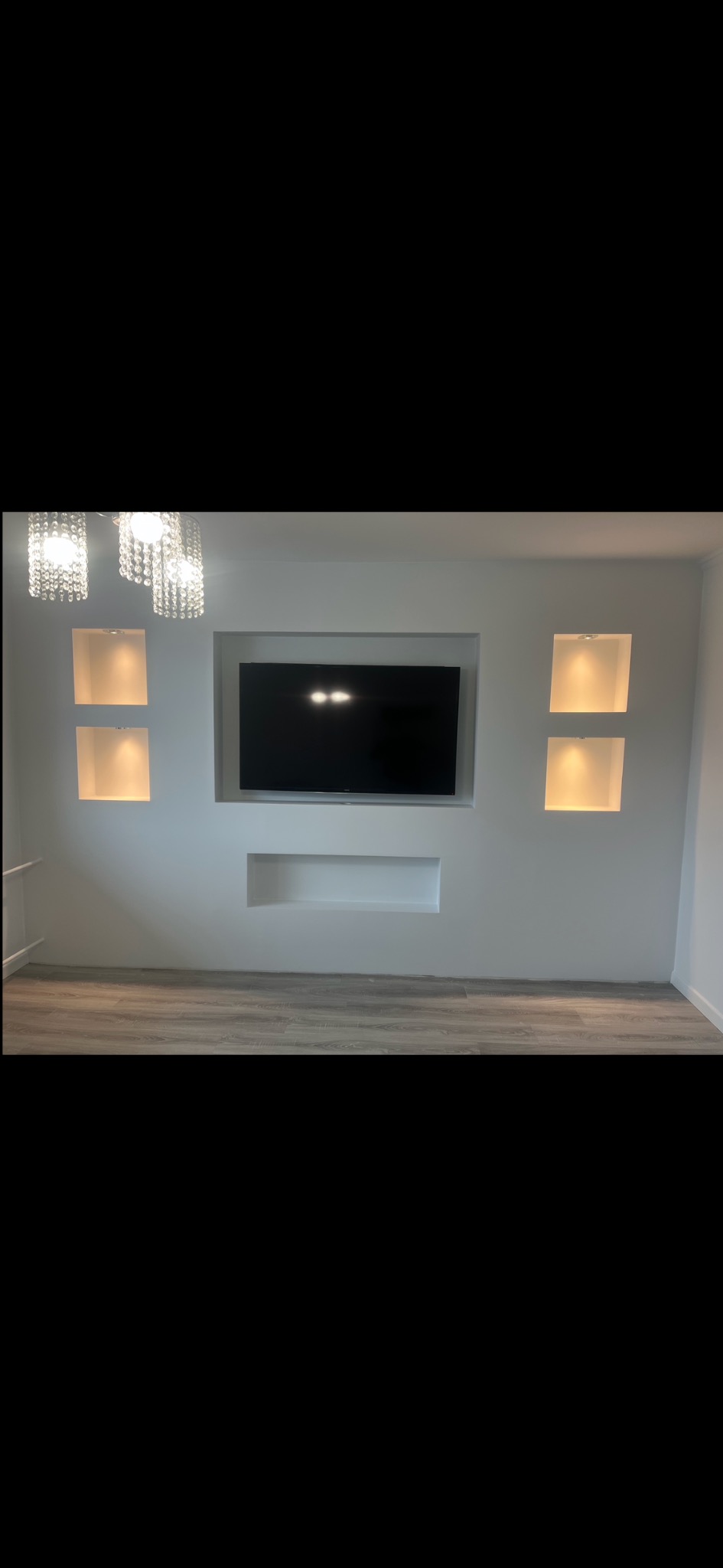 Biała ściana z wnękami podświetlonymi punktowo, w centralnej wnęce zawieszony telewizor. Minimalistyczny design wnętrza z parkietem i kryształowym żyrandolem.