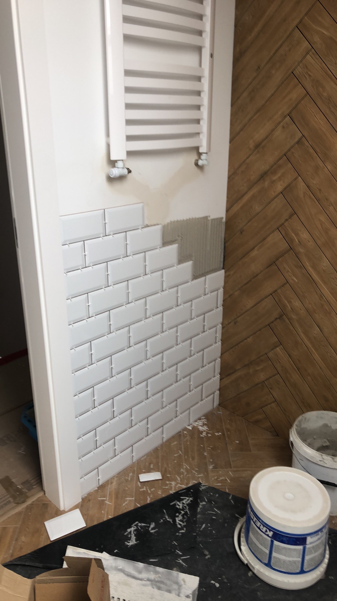 Układanie białych, prostokątnych płytek na ścianie obok drewnianej podłogi w jodełkę, widoczny grzejnik i wiaderka z klejem. Prace glazurnicze w toku.