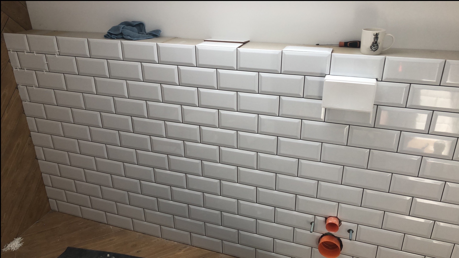 Białe, prostokątne płytki ułożone na ścianie w łazience, z widocznymi otworami na instalację wodną i krzyżykami dystansowymi. Na górze kubek i narzędzia.