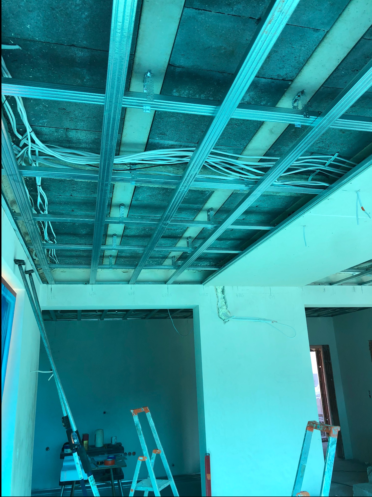 Sufit podwieszany w trakcie montażu: metalowa konstrukcja, przewody elektryczne. Widoczne drabiny i narzędzia, sufit częściowo obłożony płytami gipsowymi.