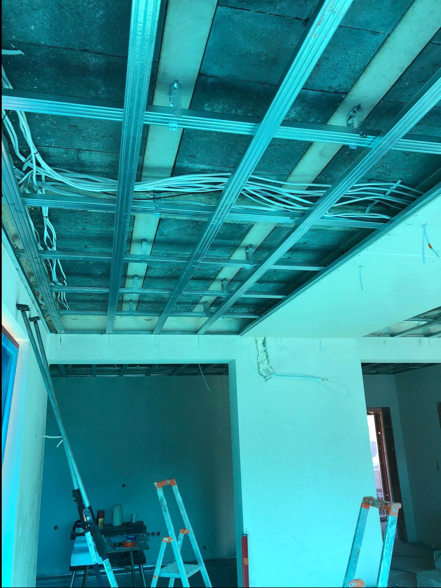 Montaż sufitu podwieszanego z płyt karton-gips, widoczne profile metalowe, instalacja elektryczna i drabina. Etap prac wykończeniowych w pomieszczeniu.