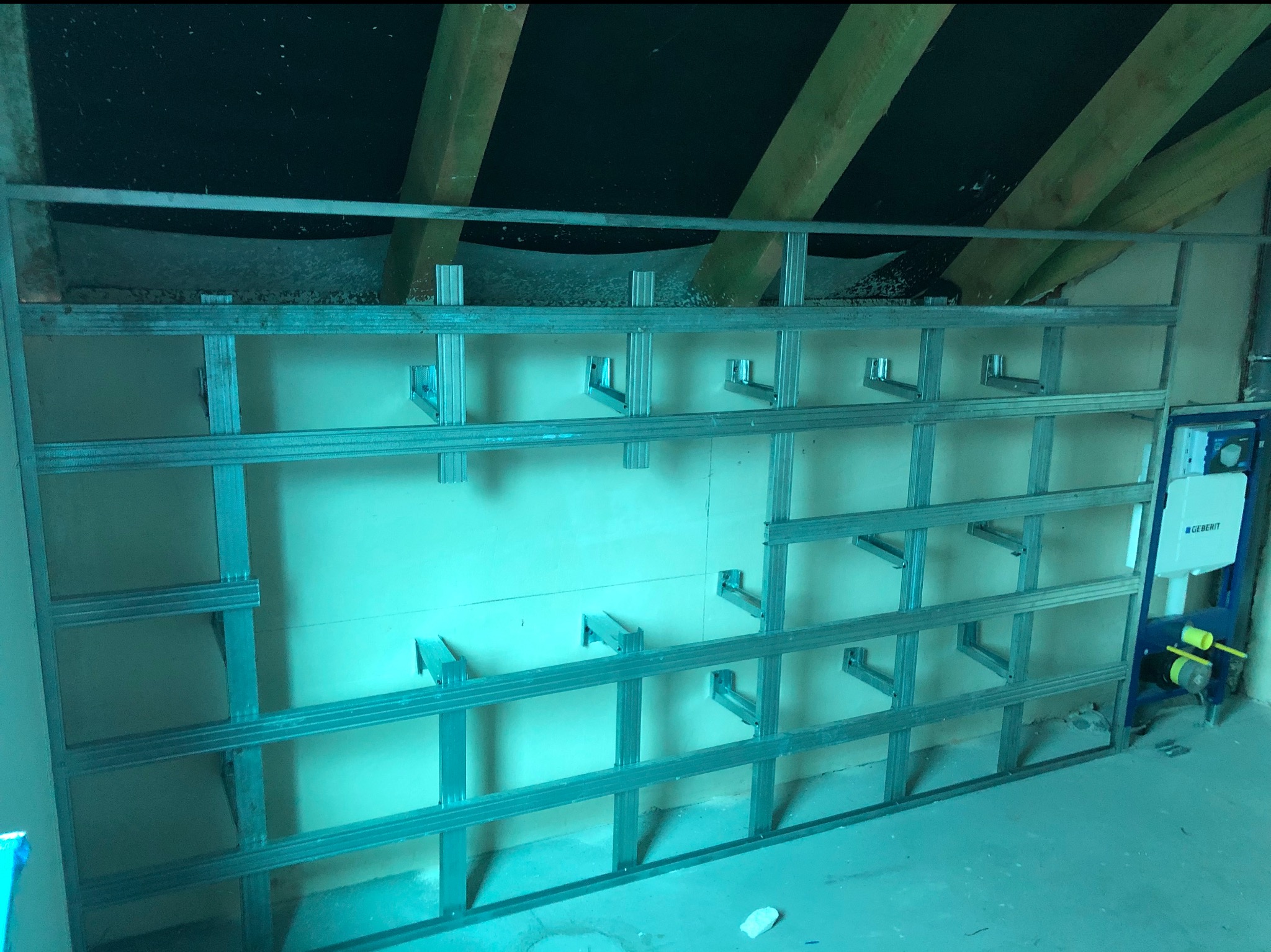 Metalowa konstrukcja stelażu pod płytę G-K na poddaszu, widoczne elementy mocujące i fragment instalacji sanitarnej Geberit.