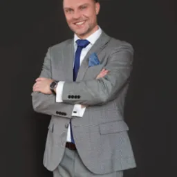 Uśmiechnięty mężczyzna w garniturze w kratę, z niebieskim krawatem i poszetką, stoi na tle ciemnoszarego tła, prezentując profesjonalny, pewny siebie wizerunek.