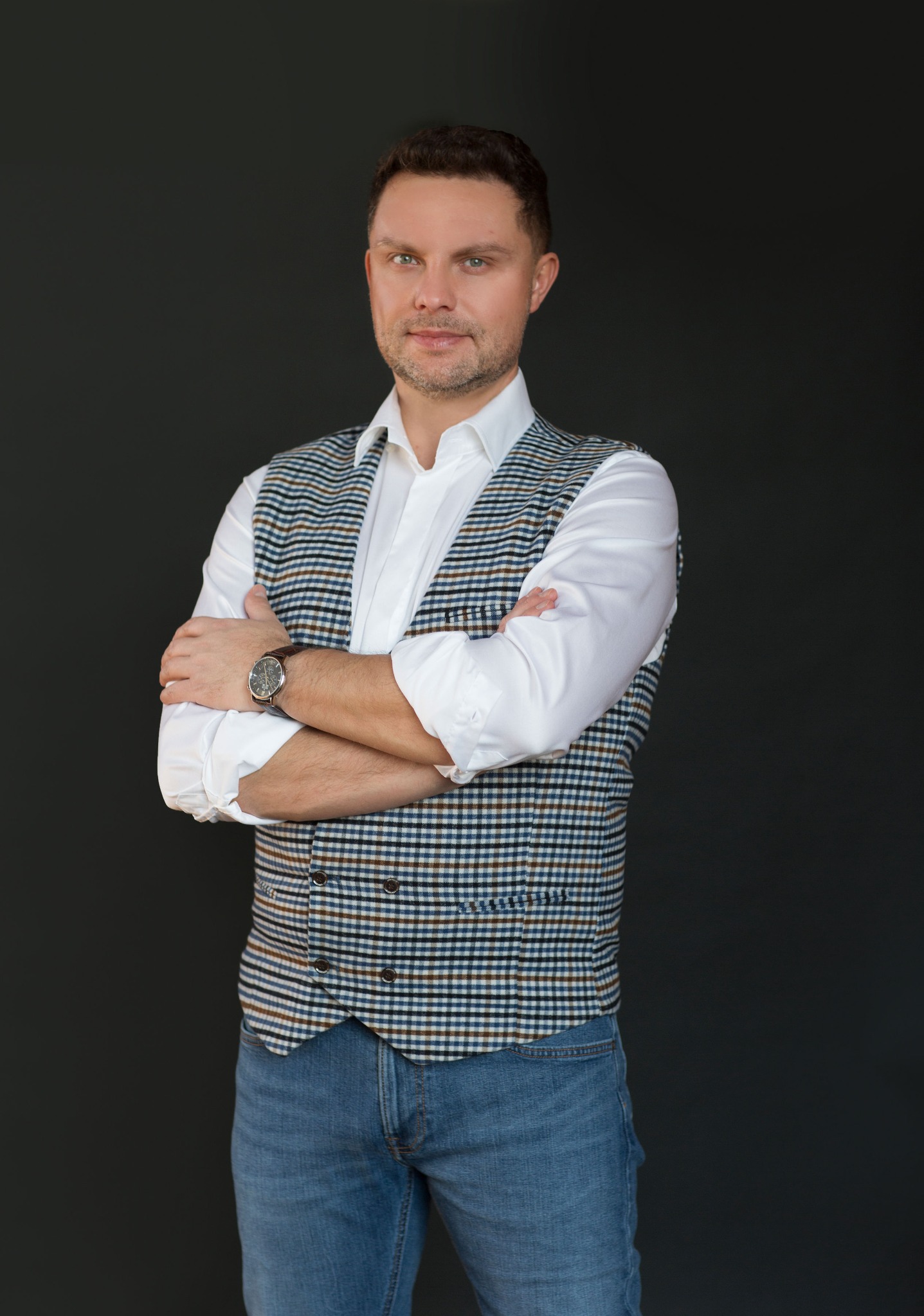 Portret mężczyzny w Lubinie, w koszuli i kamizelce w kratę, z zegarkiem na ręce, na ciemnym tle. Skrzyżowane ręce dodają pewności. Profesjonalizm.
