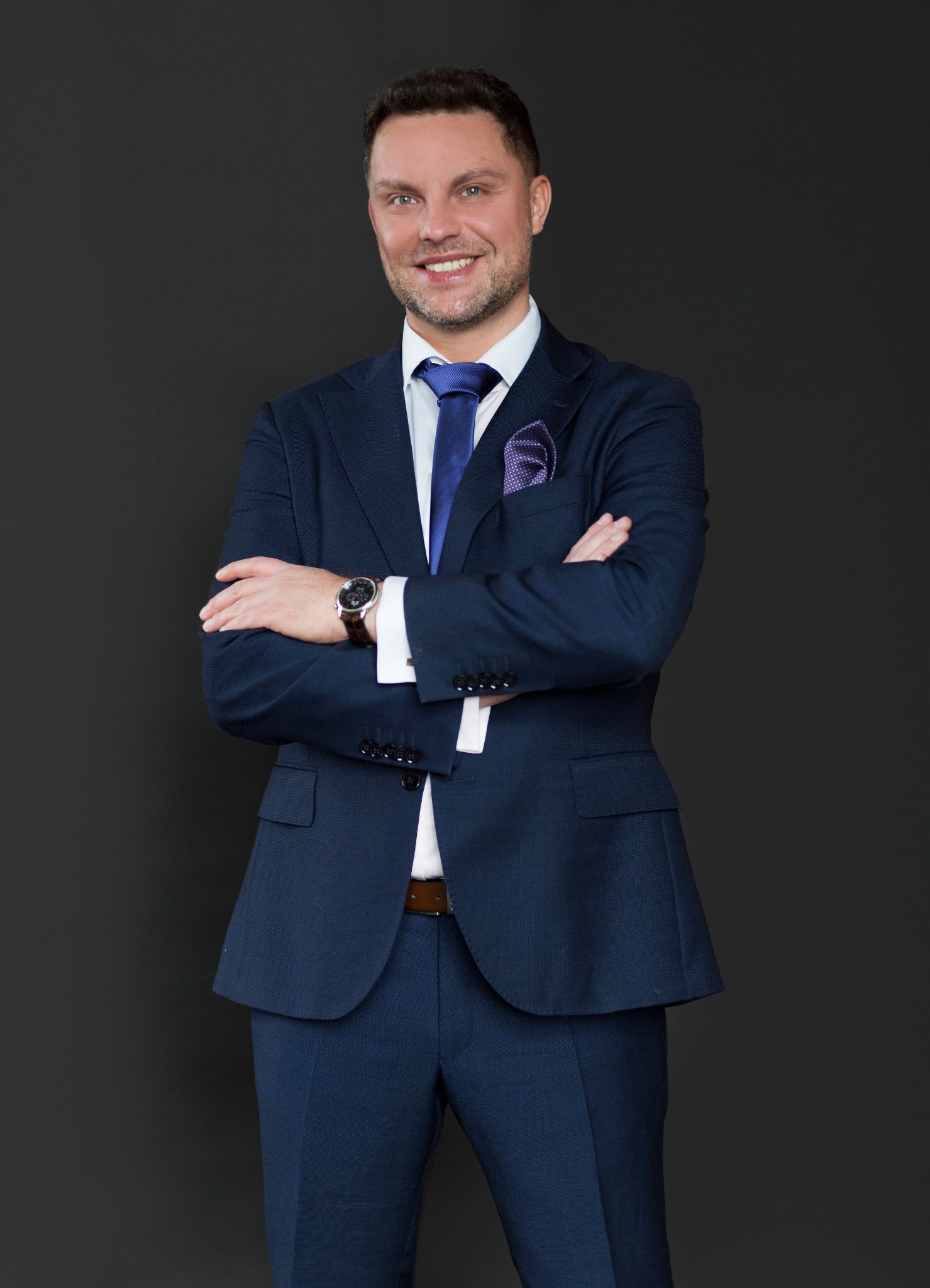 Uśmiechnięty mężczyzna w granatowym garniturze, białej koszuli i niebieskim krawacie, z zegarkiem na ręku, pozuje na ciemnoszarym tle.