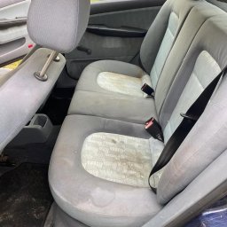 NoVel Clean - Zabrudzone tylne siedzenia samochodu przed czyszczeniem. Widoczne plamy i zanieczyszczenia na tapicerce. Fragment dywanika samochodowego z widocznym brudem. Ujęcie z otwartymi drzwiami.