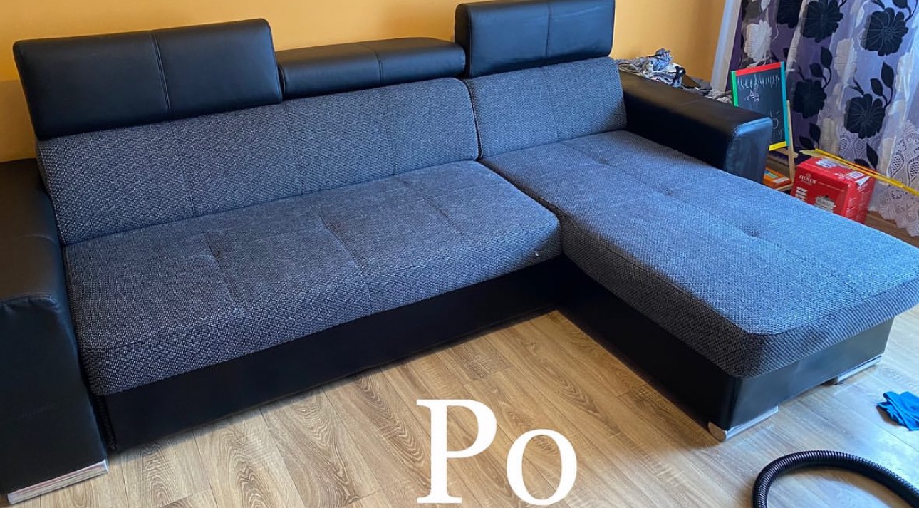 Szara kanapa narożna z czarnymi, skórzanymi elementami, widok z przodu na parkiet z napisem 'Po'. W tle firanka i przedmioty.