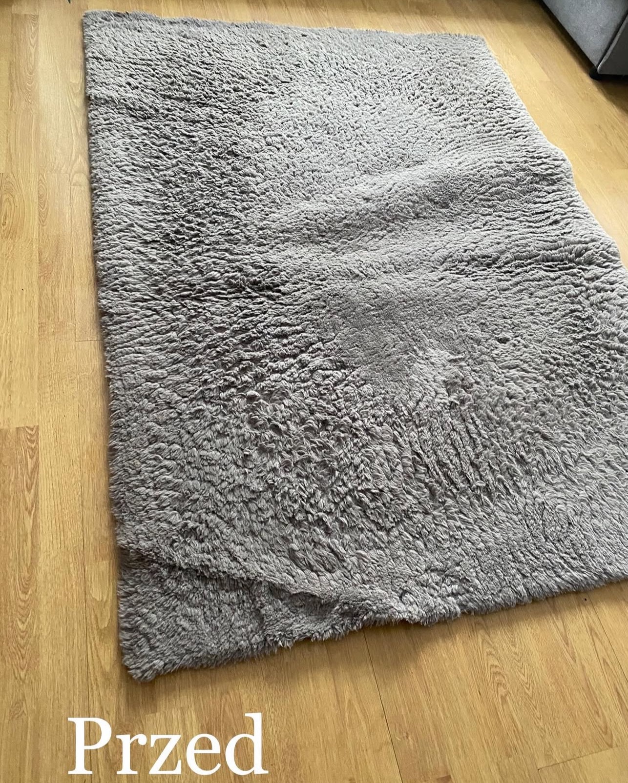 Szary, włochaty dywan na drewnianej podłodze, sfotografowany z góry. Widoczny napis 'Przed', sugerujący stan dywanu przed czyszczeniem.