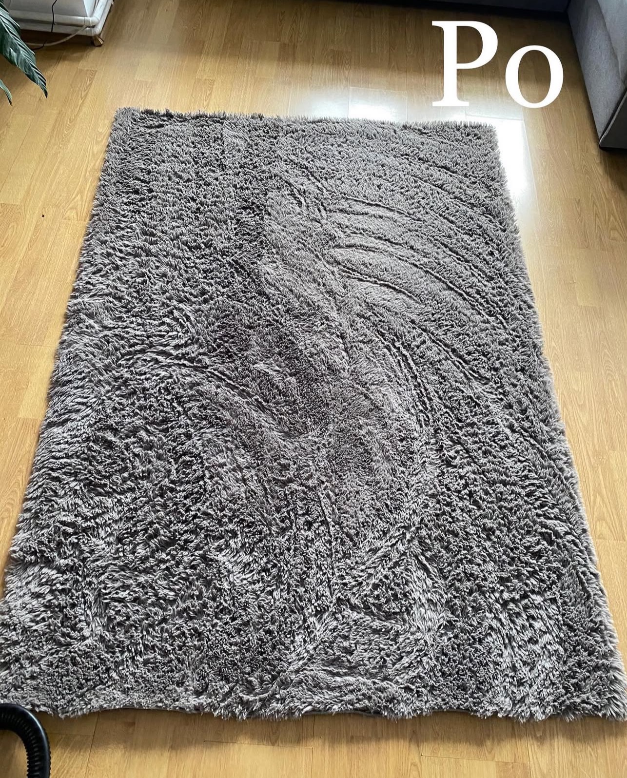 Szary, puszysty dywan typu shaggy leżący na drewnianej podłodze, oznaczony napisem 'Po', sugerujący efekt po praniu.