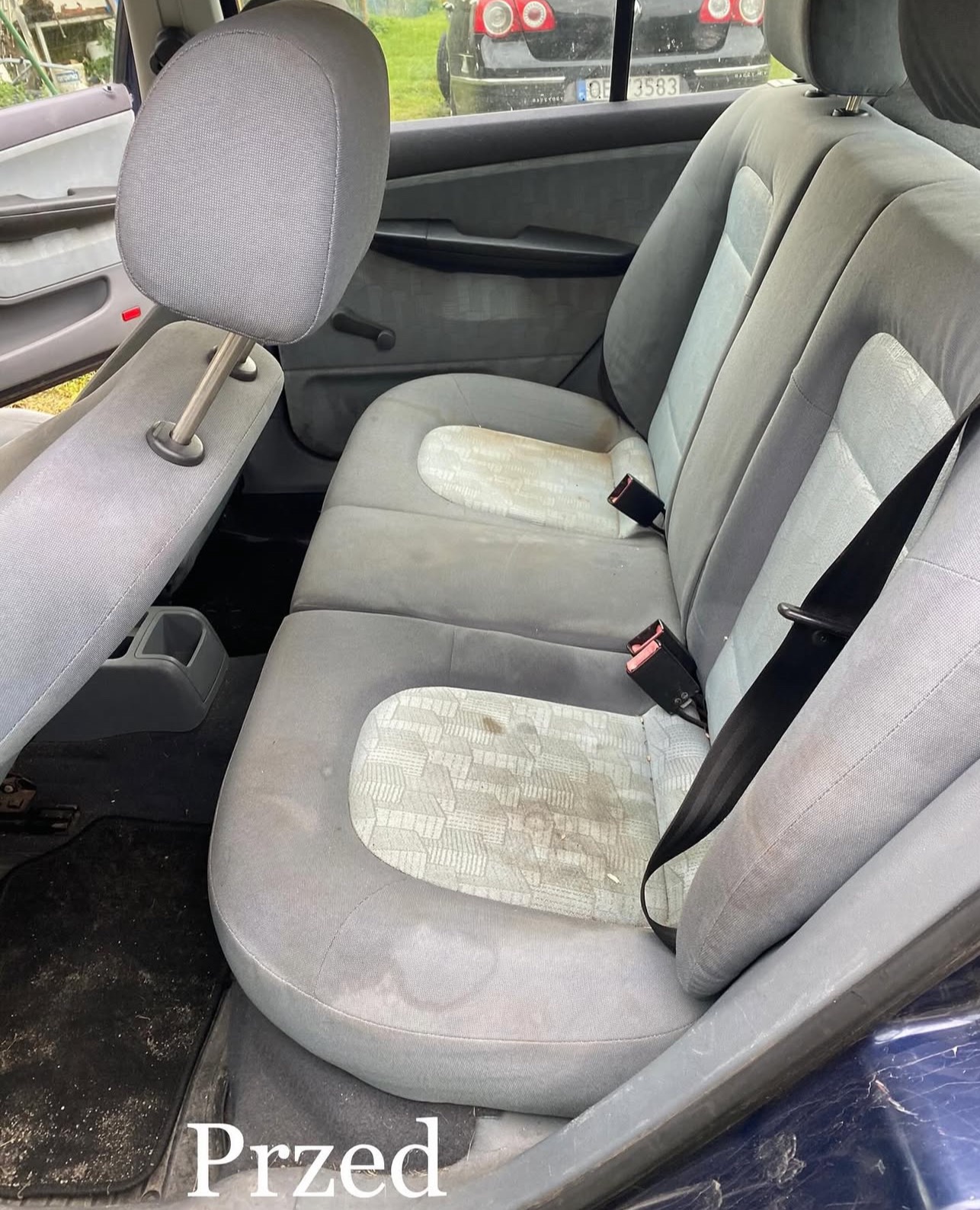 Zabrudzone tylne siedzenia samochodu przed czyszczeniem. Widoczne plamy i zanieczyszczenia na tapicerce. Fragment dywanika samochodowego z widocznym brudem. Ujęcie z otwartymi drzwiami.