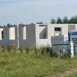 Wznoszenie ścian domu z betonu komórkowego. Widoczny pracownik w oknie, palety z bloczkami i przenośna toaleta. Tło stanowi zieleń drzew i krzewów.