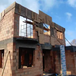 Surowy stan domu z czerwonej cegły, widoczne otwory okienne i przygotowanie do dachu, na tle błękitnego nieba z chmurami. Etap budowy z widocznymi elementami konstrukcyjnymi.