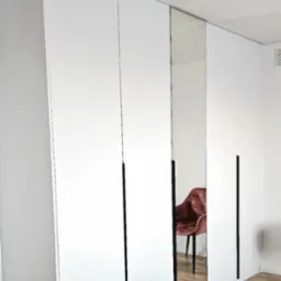 Biała szafa wnękowa z lustrzanymi drzwiami i czarnymi uchwytami, odbijająca krzesło w pokoju, widok z dołu, częściowo widoczne łóżko.