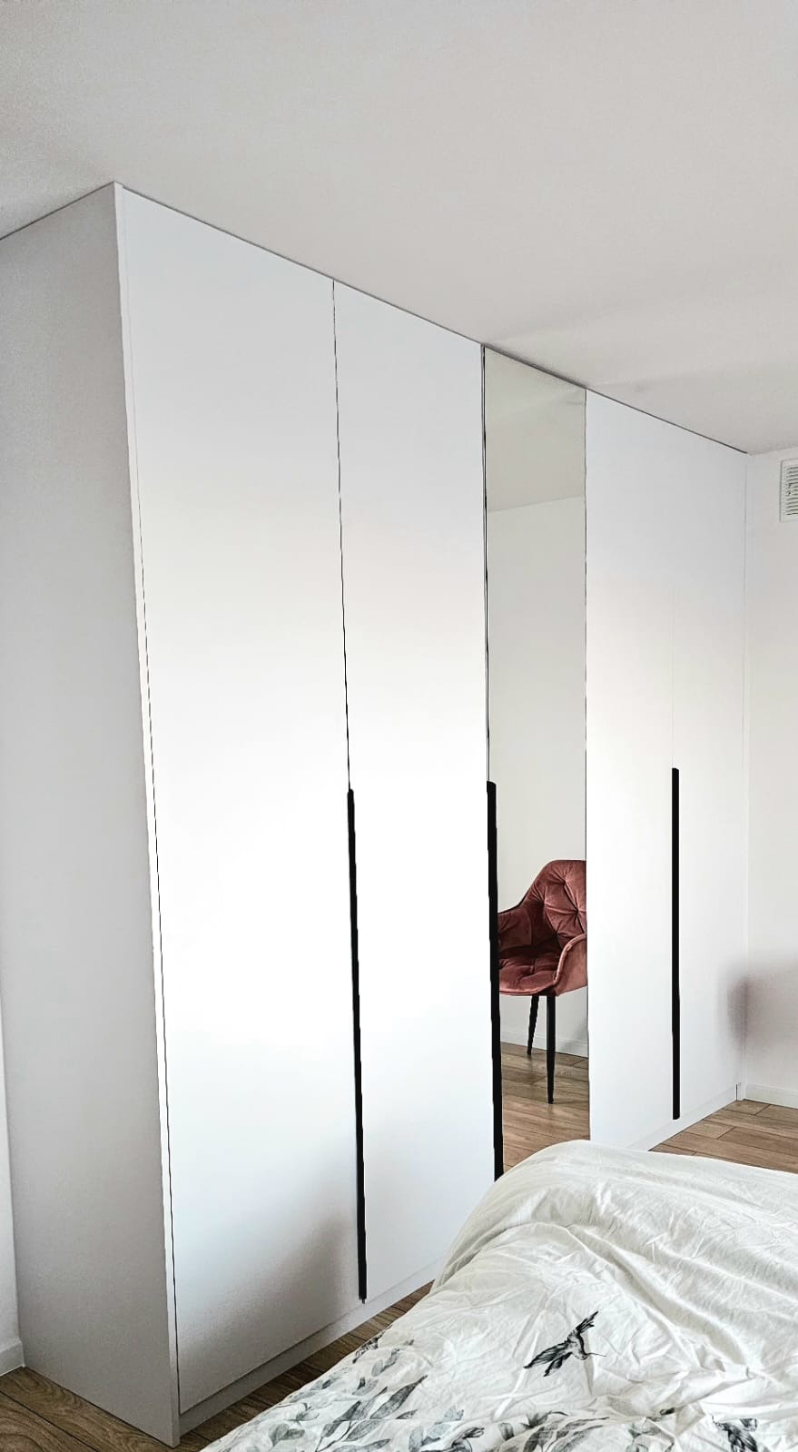 Biała szafa wnękowa z lustrzanymi drzwiami i czarnymi uchwytami, odbijająca krzesło w pokoju, widok z dołu, częściowo widoczne łóżko.