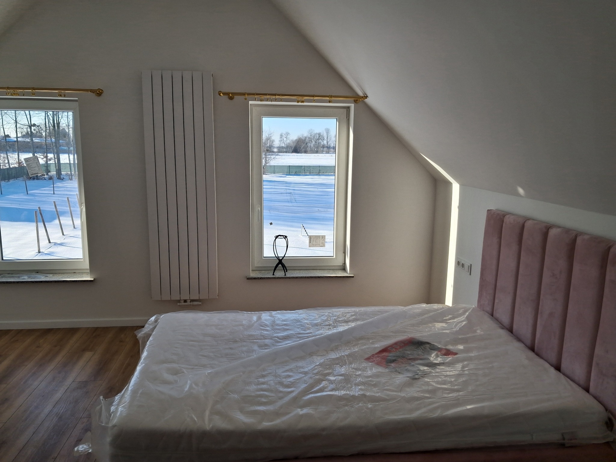 Jasne poddasze z łóżkiem zabezpieczonym folią, grzejnikiem i oknami z widokiem na zimowy krajobraz. Parkiet na podłodze, skosy sufitowe.