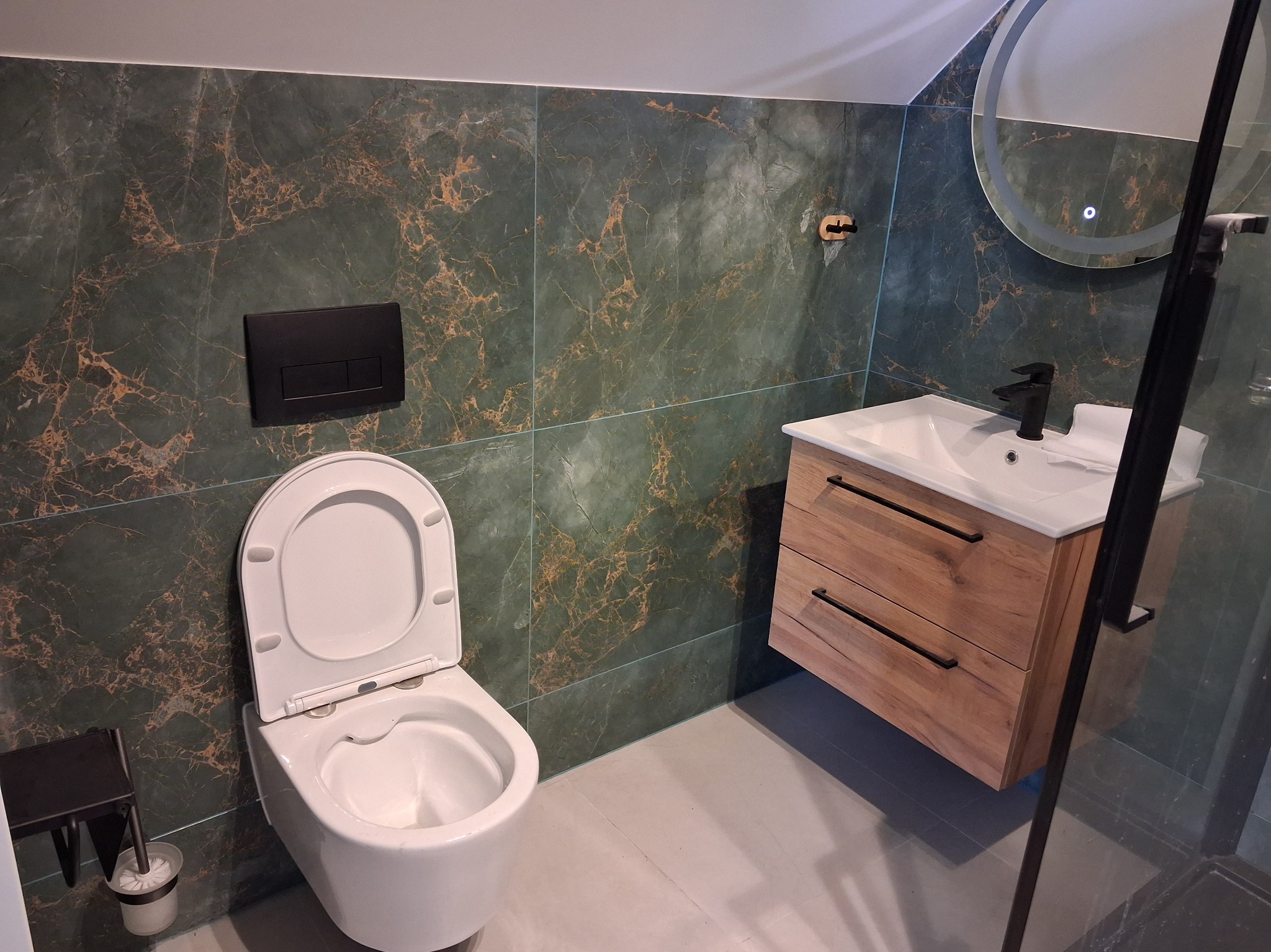 Nowoczesna łazienka z zielonymi płytkami imitującymi marmur, drewnianą szafką pod umywalką i okrągłym lustrem z podświetleniem LED. Widoczny fragment kabiny prysznicowej.