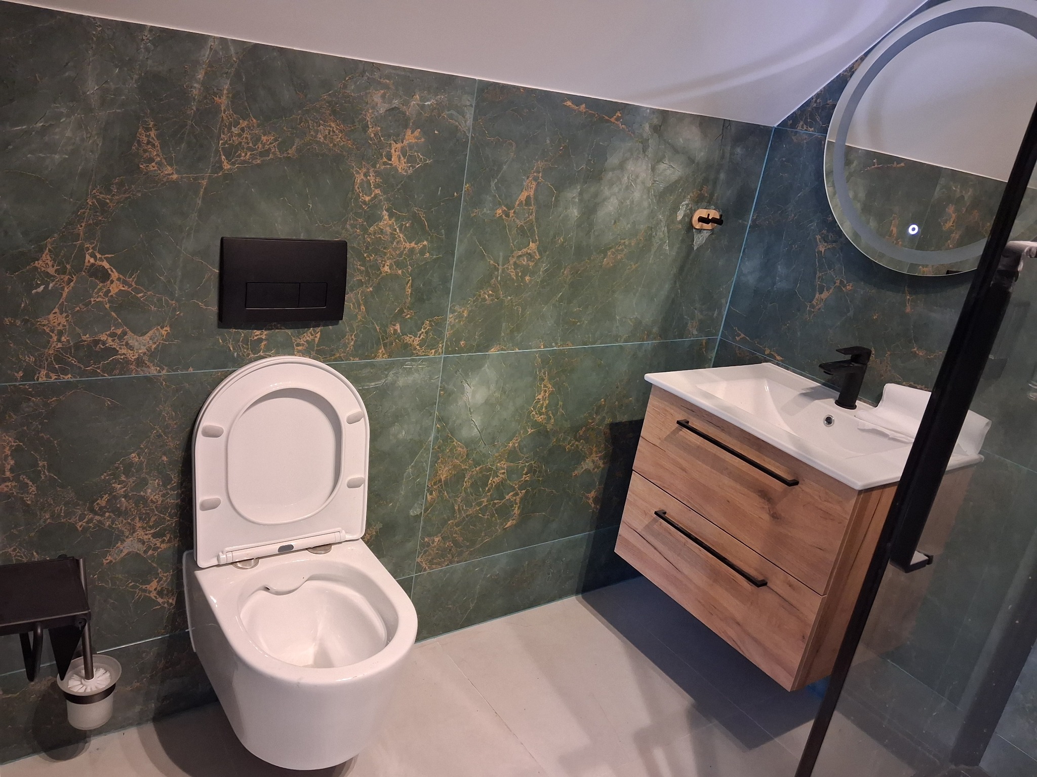 Nowoczesna łazienka z zielonymi płytkami imitującymi marmur, drewnianą szafką pod umywalką i okrągłym lustrem z podświetleniem LED. Widoczna toaleta i czarne akcenty.