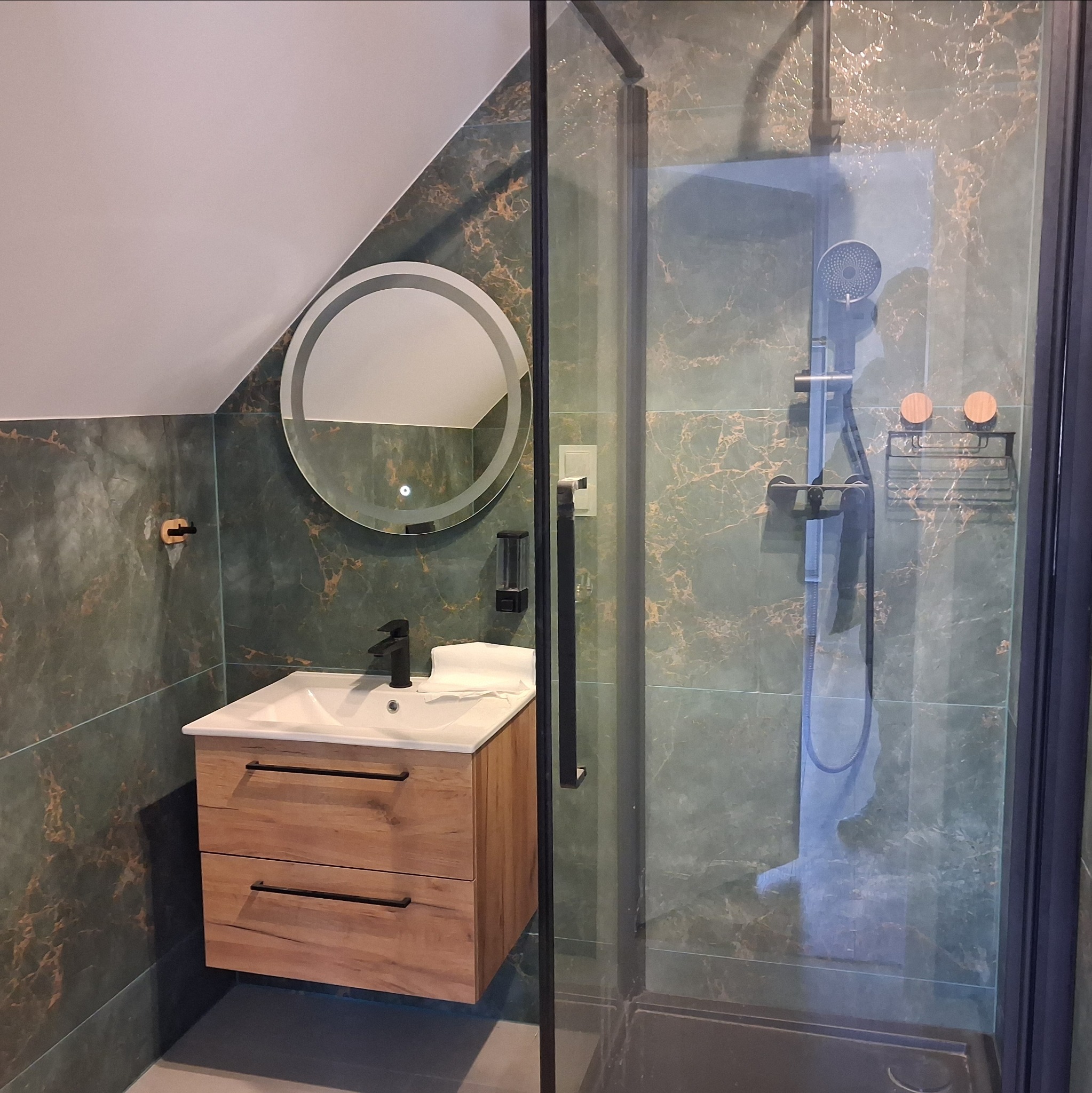 Nowoczesna łazienka na poddaszu z kabiną prysznicową, umywalką z drewnianą szafką i okrągłym lustrem z podświetleniem. Marmurowe płytki w odcieniach zieleni i złota.