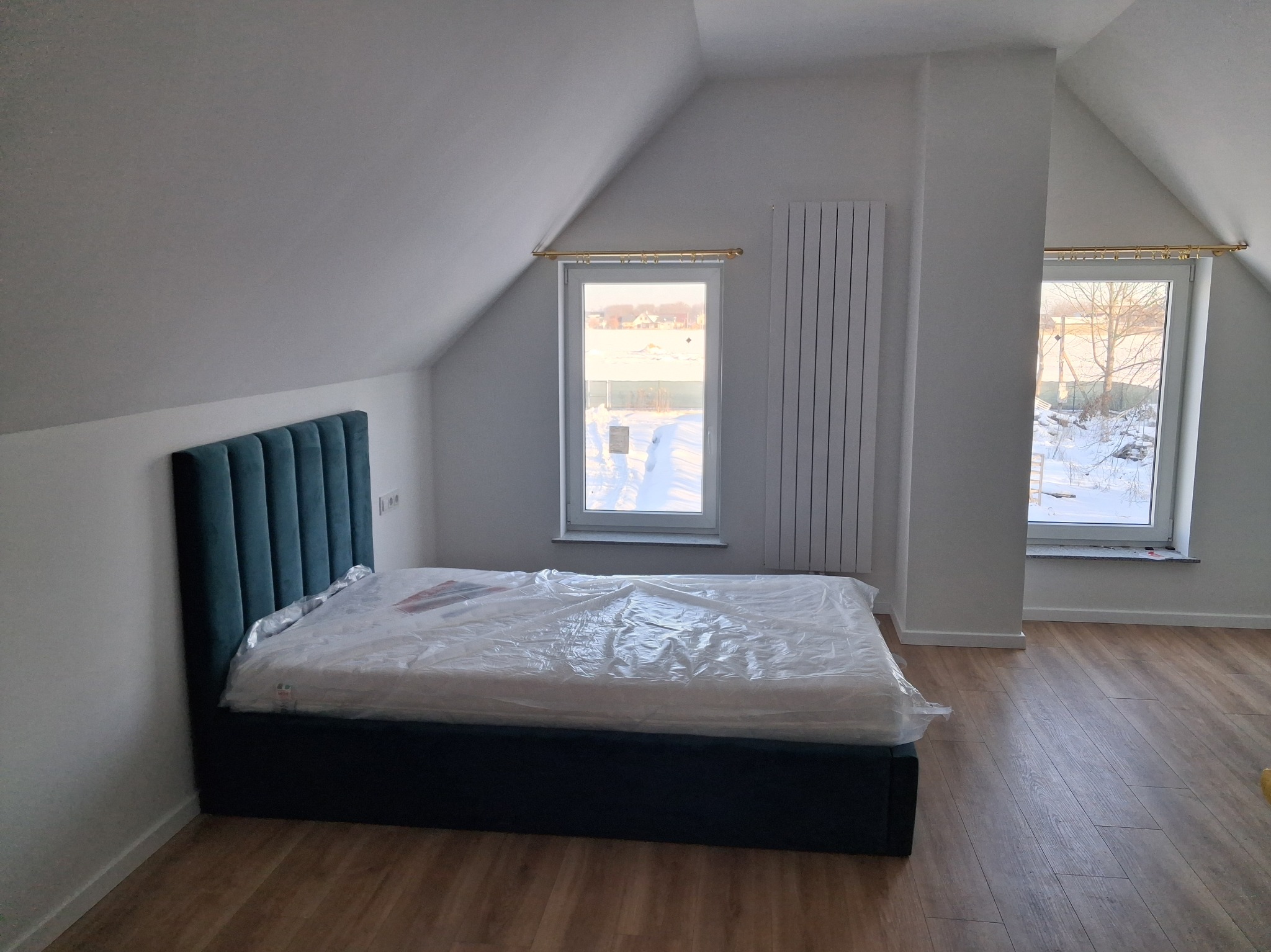 Nowe łóżko z zielonym zagłówkiem w pokoju na poddaszu, jeszcze owinięte folią ochronną. Widoczne okna z widokiem na zimowy krajobraz i drewniana podłoga.