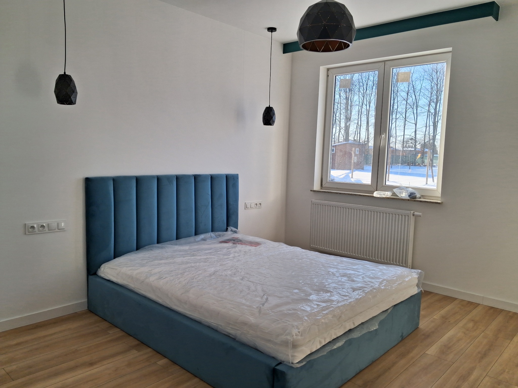 Wykończona sypialnia w Pruszkowie: błękitne łóżko z wezgłowiem, drewniana podłoga, białe ściany i okno z widokiem na zimowy krajobraz. Nowoczesne lampy wiszące.