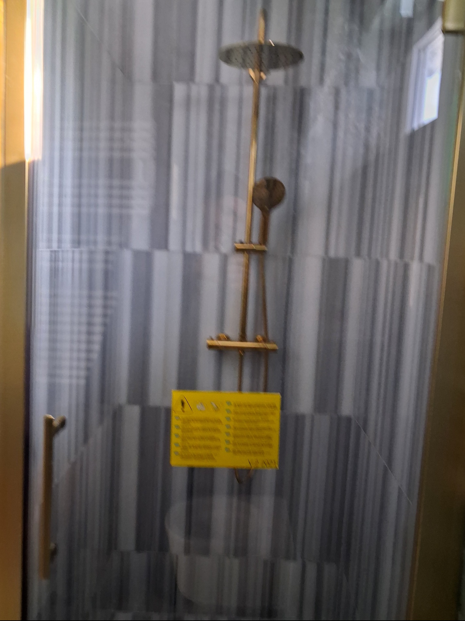 Kabina prysznicowa z deszczownicą i szarymi płytkami w paski. Na szybie żółta naklejka informacyjna. Złote wykończenia armatury i ramy kabiny.