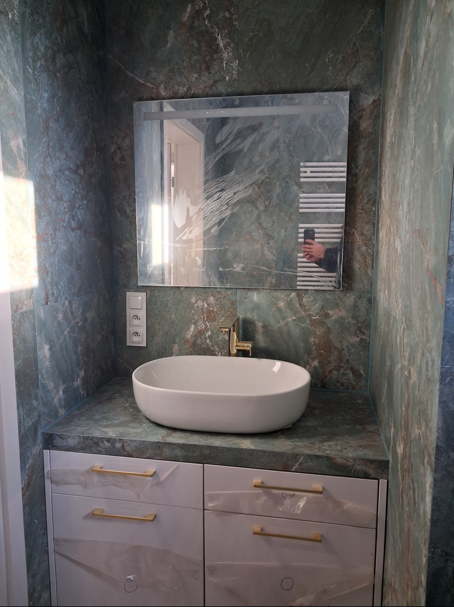 Wykończona łazienka z umywalką nablatową, złotą armaturą i marmurowymi płytkami w odcieniach zieleni. W lustrze odbija się grzejnik drabinkowy i osoba robiąca zdjęcie.